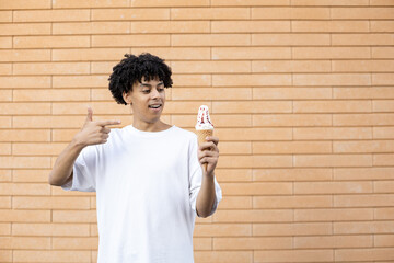Fototapeta premium Dessert, summer, sweets and people concept - Smiling, curly-haired African-American guy holding an ice cream cone and pointing his finger at it, wearing a white T-shirt on a brick wall background