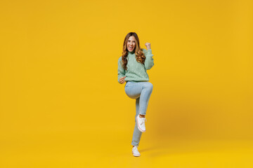 Fototapeta na wymiar Full body young overjoyed excited woman 30s wearing green knitted sweater do winner gesture clench fist raise up leg isolated on plain yellow color background studio portrait People lifestyle concept