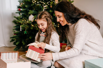 Excited mother and daughter sitting at home near beautiful decorated Christmas tree and enjoying opening their presents. Single solo parenting holidays. Family happiness. Selective focus