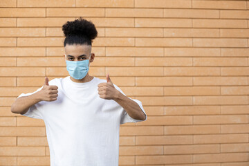 Health, pandemic, safety and people concept - A young African-American guy showing thumbs up with both hands in a protective mask from viruses dressed in a white T-shirt on a brick wall background