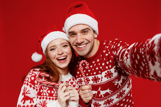 Close up fun young couple friends two man woman 20s in sweater hat do selfie shot pov on mobile phone hold candy cane lollipop isolated on plain red background Happy New Year 2022 celebration concept.
