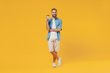 Full body young smiling happy man in blue shirt white t-shirt hold takeaway delivery craft paper brown cup coffee to go isolated on plain yellow background studio portrait. People lifestyle concept.