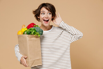 Young smiling vegetarian woman in casual clothes hold paper bag with vegetables after shopping in...