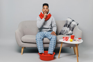 Young african man in grey sweater scarf sit on sofa say hush be quiet with finger on lips shhh...