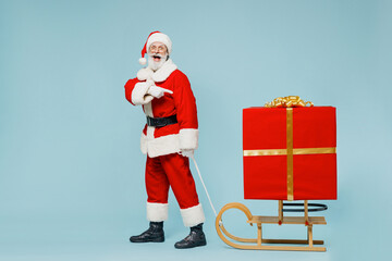 Full body old Santa Claus man 50s in Christmas hat red suit clothes pull sled with gift point on big box isolated on plain blue background studio. Happy New Year 2022 merry ho x-mas holiday concept.