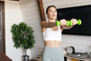 Fototapeta na wymiar Fitness asian girl doing workout at home in living room, lifting dumbbells, wearing activewear for sport activities