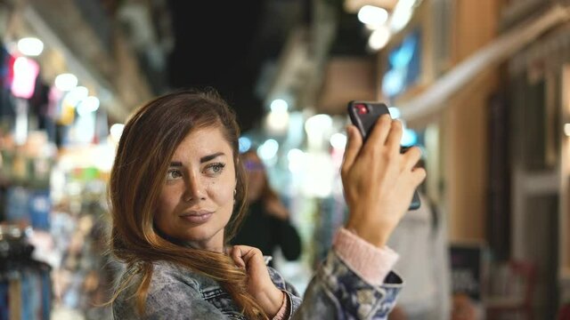 Young adult lady with iphone takes selfie, making photos of herself. Traveling, tourism to Athens in Greece. Spending time on pictures with phone in dusk evening time. Having casual fun in Greek city.