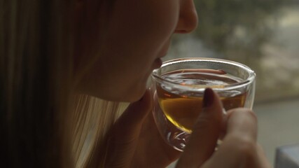 A young girl is sitting near the window and holding a glass mug with warm tea blowing cool air into her cup