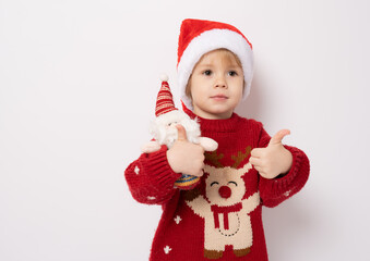 Little boy wearing santa hat holding santa claus toy with thumb up standing over white background.