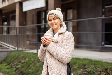 Young pretty woman, 28 years old, wearing knitted hat, coat made of faux beige fur, warming up on winter day holding recyclable cup of coffee or tea, lifestyle, street style, city life. - obrazy, fototapety, plakaty