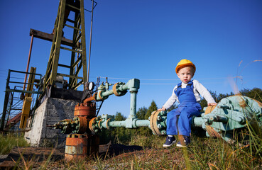 Cute child in construction helmet sitting on pipe of petroleum pump jack under beautiful blue sky....