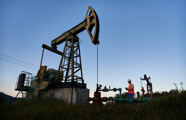 Mechanic in work vest and construction helmet standing near pipeline, to the left of pump jack and...