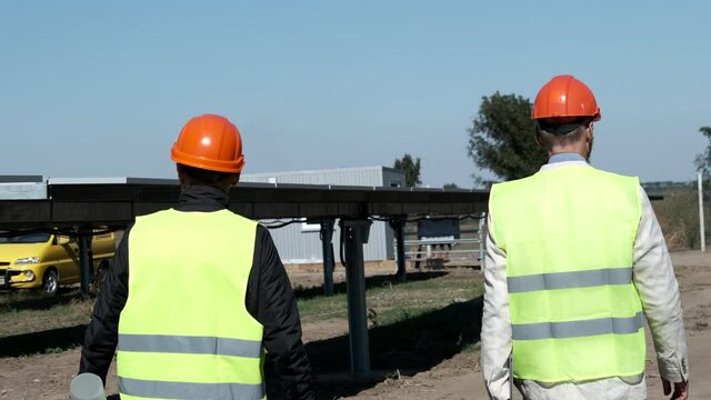 Solar and wind clean power. Specialists of maintenance in orange helmets walk past sun collecting panels at construction site backside view