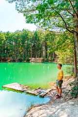 Fototapeta na wymiar A blond man stands in front of an azure mountain lake in the forest in summer