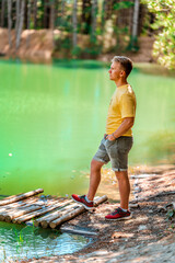 A blond man stands in front of an azure mountain lake in the forest in summer