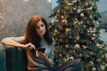 A woman sits thoughtfully with a phone in her hand while sitting on the couch near the Christmas tree.