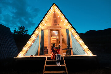 Woman drinking tea on the porch of a wooden lodge with lights of garlands in the evening. The concept of glamping and renting a chalet for weekend - obrazy, fototapety, plakaty