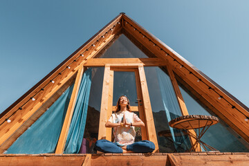 Calm woman meets a new day in a yoga pose on the porch of a cute little wooden house in the...