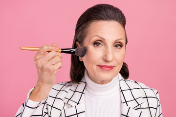 Portrait of mature lovely charming lady applying blush on cheeks natural makeup isolated on pink color background
