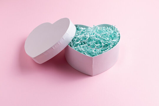 White Heart Shape Gift Box Full Of Blue Crinkle Cut Paper Shred Filler On Pastel Pink Background. Shot At Angle.