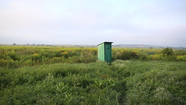 Old wooden toilet with a carved window with the shape of a heart cut out on the door, in open field. Vintage WC. An outdoor rustic green restroom in a field landscape of flowers and grass at sunrise.