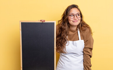 hispanic pretty woman shrugging, feeling confused and uncertain. blackboard copy space concept