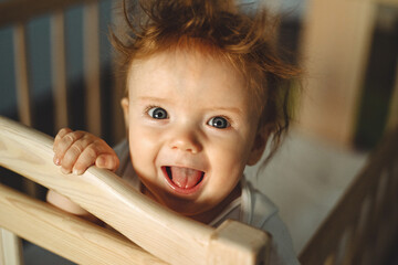 Funny shaggy baby with red hair looks out of the crib and laughs.
