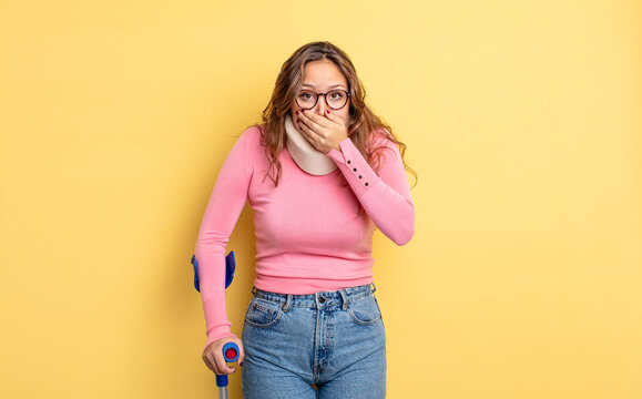 hispanic pretty woman covering mouth with hands with a shocked. crutch and collar accident