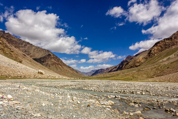 The wide river bed of the Kargyak river with the mountains of the Zanskar range and a blue sky on the Darcha Padum trekking route in the Indian Himalaya.