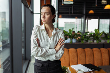 Fototapeta premium Stylish businesswoman in white shirt standing with crossed arms and pondering while looking away in cafe 