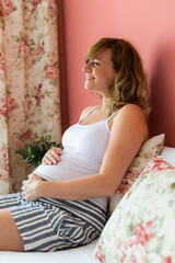 A smiling pregnant woman is sitting in bed, looking trough the window.