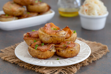 Halved potatoes baked with onion and chopped bacon. Portion served on a white plate with sauerkraut salad. Dark background, selective focus. 
