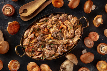 Beef stroganoff, mushroom and meat ragout in a creamy sauce, in a pan with ingredients, overhead flat lay shot on a dark background