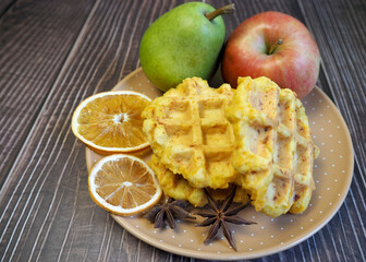 Belgian gluten-free pumpkin waffles lie on a gray plate with a dry slice of lemon and an apple and pear on a wooden table. side view