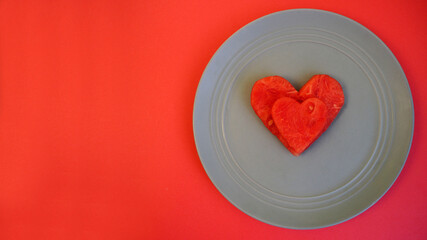 two red slices of watermelon in the shape of a heart lie side by side on a gray plate on a red background