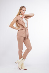 High fashion photo of a beautiful elegant young woman in a pretty beige suit, pants, trousers, vest, boots posing over white, soft gray background. Studio Shot, portrait. Blonde