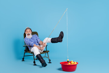 Full size photo of happy grey haired positive cheerful old man enjoy fish hold rod sit chair isolated on blue color background