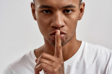 Fototapeta na wymiar Portrait of dark skinned young guy showing shh gesture, keeping a secret, posing isolated over white background
