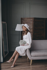 girl in a white dress and a white hat with wide brim poses while sitting on a sofa with a bouquet of white flowers.