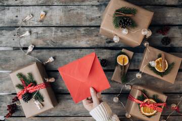 Top view woman hands in sweater holding blank red envelope . Flat lay on wooden background with Christmas garland, presents and natural decorations. Top view mock up and copy space for text.