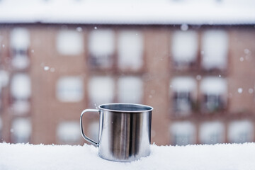 Obraz na płótnie Canvas metallic cup of hot drink, tea or coffee, on terrace during snow in winter, neighborhood background. Snow in the city. Nobody