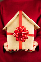 Young woman in red sweeter holding a house wrapped in eco rustic beige recycled brown paper and tied with a red ribbon with a bow. Christmas, New Year, Birthday, Valentine's Day concept. Close-up