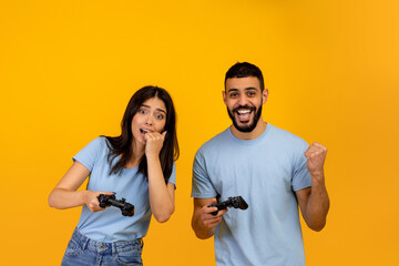 Emotions of win and loss in online game. Playful arab guy rejoicing success near upset woman, yellow background © Prostock-studio
