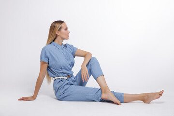 High fashion photo of a beautiful elegant young woman in a pretty blue jumpsuit, barefoot posing over white, soft gray background. Studio Shot, portrait. The model is sitting in the background. Blonde