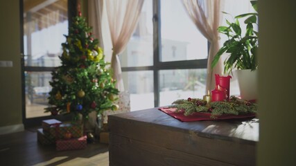 An evergreen wreath with two red candles and one golden candle. In the background, a decorated Christmas tree. Christmas traditions and customs of Christians on the eve of the holiday.