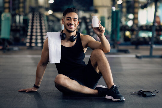 Young Muscular Arab Man Holding Container With Supplement Pills, Posing At Gym