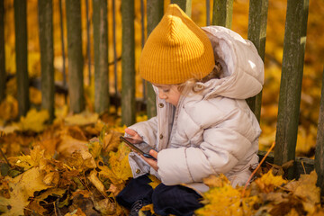 Stylish child sits on the ground in the leaves outdoors, holding a smartphone in his hands, watching cartoons.