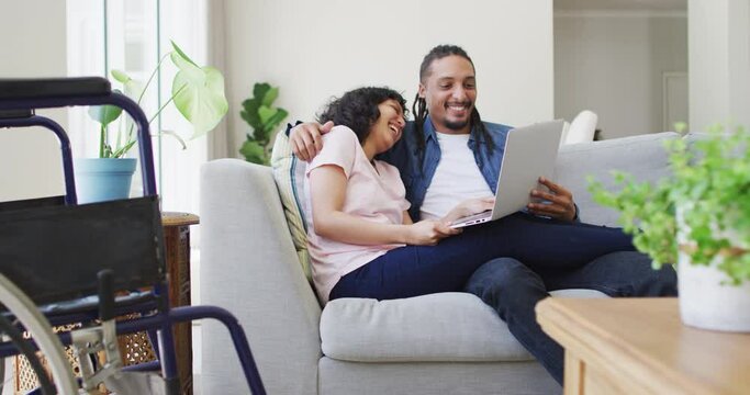 Happy biracial couple in living room using laptop and embracing, with her wheelchair beside couch
