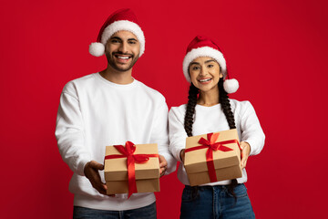 Cheerful Middle Eastern Couple In Santa Hats Giving Present Boxes At Camera
