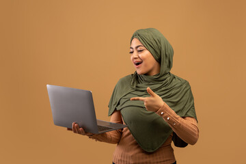 Excited muslim woman in hijab holding laptop and pointing at pc, looking at screen with excitement,...